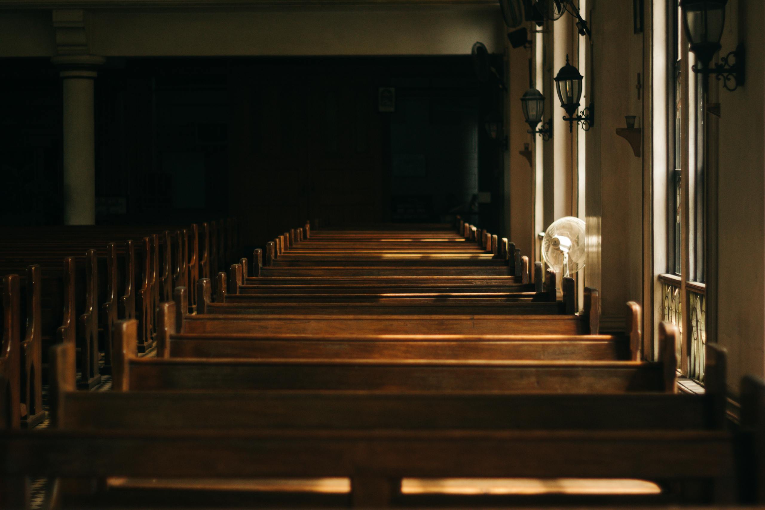 Empty wooden pews inside a dimly lit church sanctuary, with sunlight filtering through windows on the right side. The peaceful, undisturbed setting reflects a quiet moment, relevant for illustrating the protection and coverage offered by church insurance quotes.