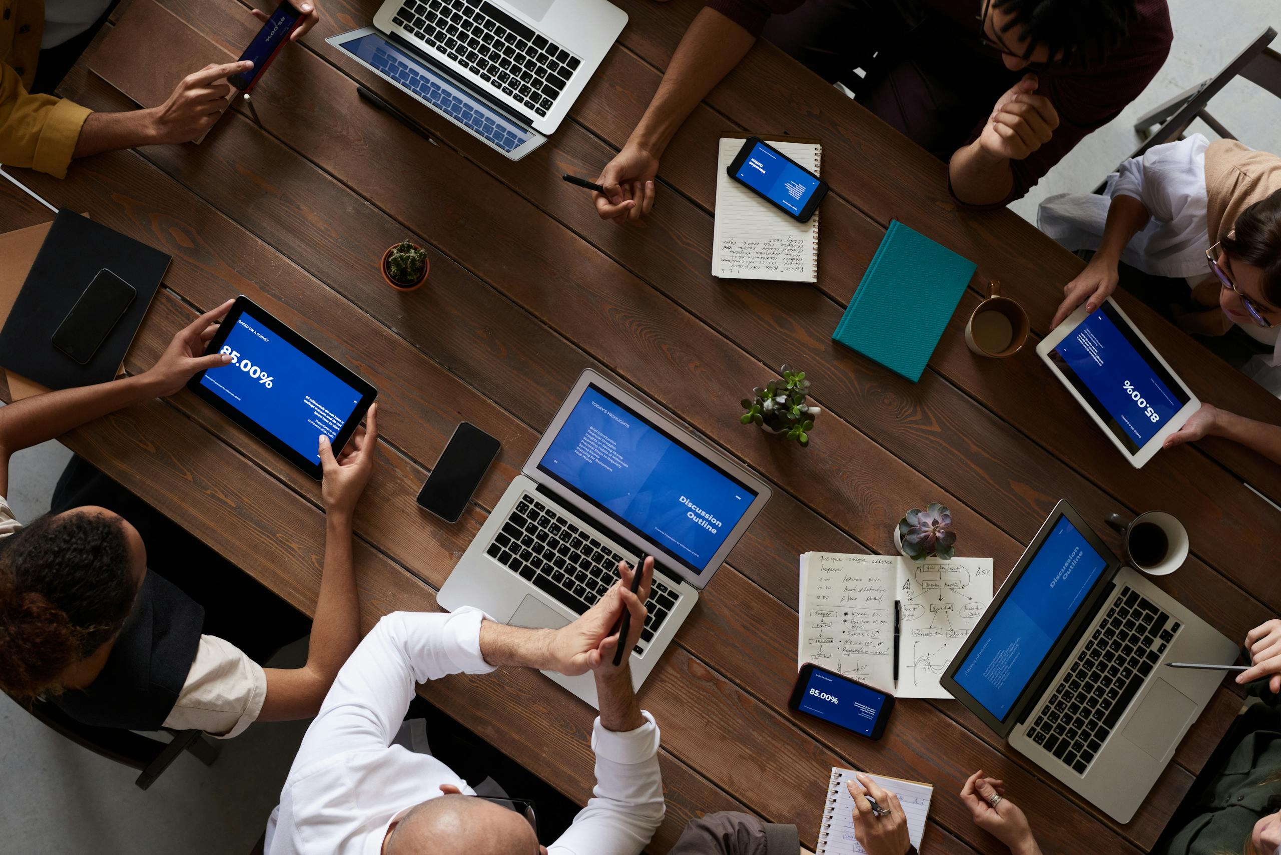 Overhead view of a diverse group of people gathered around a wooden table, working collaboratively on laptops, tablets, and phones. This image reflects teamwork and professional settings, making it a strong visual for content related to GVRA insurance compliance.